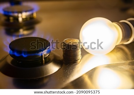 Light bulb on next to lit gas cooker, with coins next to it. Energy and gas costs, cost increases. Royalty-Free Stock Photo #2090095375