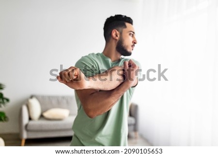 Athletic young Arab guy stretching arm muscles, doing warmup exercises at home, selective focus. Millennial middle Eastern man having domestic training, working out indoors Royalty-Free Stock Photo #2090105653