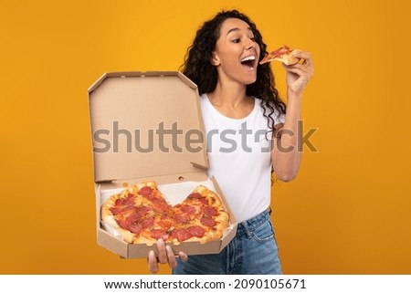 Excited Latin Young Lady Enjoying Pizza Holding And Biting Tasty Slice Posing With Carton Box Over Yellow Orange Studio Background. Junk Food Lover Eating Italian Pizza. Unhealthy Nutrition Cheat Meal