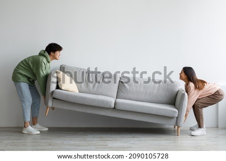 Happy young Asian couple moving sofa in living room, replacing furniture at home, copy space. Millennial husband and wife making rearrangement in their apartment, side view Royalty-Free Stock Photo #2090105728