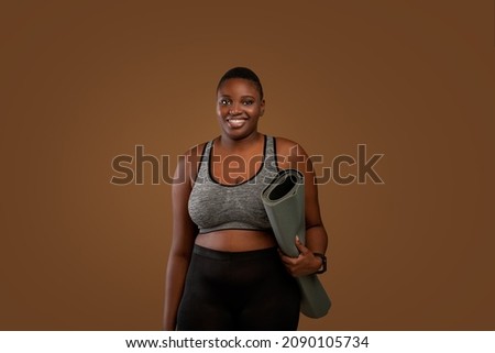 Ready For Training. Portrait of African American chubby woman in bra top and leggings going to gym, holding carrying roll mat standing isolated over brown studio background, looking posing at camera