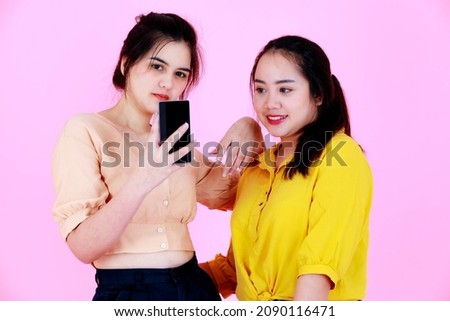 Studio shot of two Asian chubby and slim braces teeth ponytail hair female friends or sisters model in casual wear crop top holding smartphone smiling taking selfie photo together on pink background.