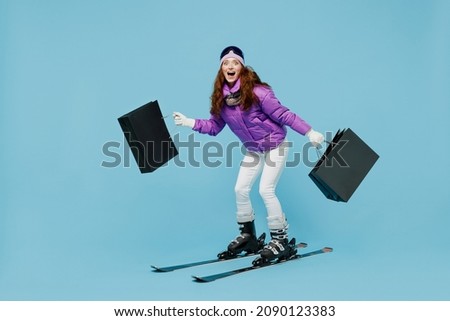 Full size amazed skier woman in warm purple padded windbreaker jacket ski goggles mask spend weekend in mountains holding package bags with purchases shopping isolated on plain blue background studio