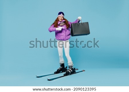 Full body skier woman in warm purple padded jacket ski goggles mask point on spend weekend in mountains holding package bags with purchases after shopping isolated on plain blue background studio.