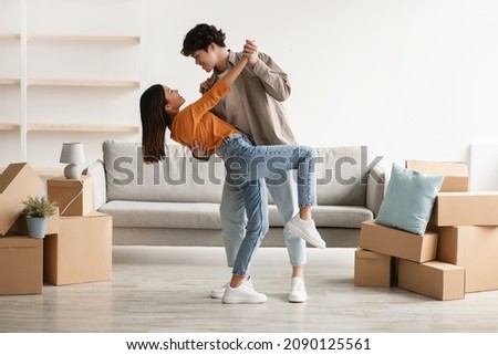 Affectionate young Asian couple dancing in their house among cardboard boxes on moving day, full length. Happy millennial spouses having fun while relocating to new property, copy space