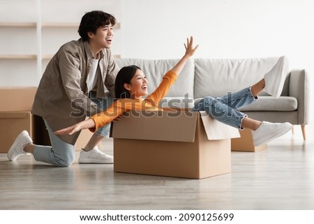 Playful young Asian couple having fun in their home on relocation day. Millennial woman and her boyfriend playing together with carton box while moving to new apartment Royalty-Free Stock Photo #2090125699