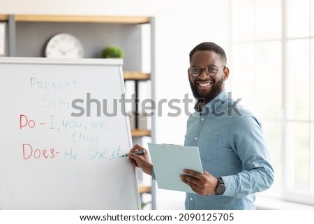 Smiling millennial african american bearded guy teacher in glasses stands near blackboard and teaches english in home interior. Remote tutoring, social distance study and knowledge during covid-19 Royalty-Free Stock Photo #2090125705