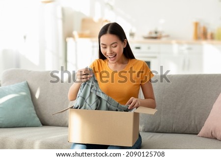 Quick purchases in online store. Satisfied asian woman opening carton box sitting on sofa in living room at home, copy space. Happy female customer taking clothing from container Royalty-Free Stock Photo #2090125708