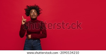 Pointing up. Flyer with image of beautiful dark skinned young girl in warm outfit standing isolated on dark red background. Concept of emotions, facial expression, youth, sales. Copy space for ad.