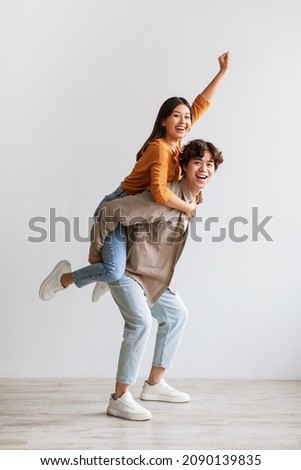 Handsome young Asian man giving piggyback ride to his girlfriend against white studio wall, full length portrait. Millennial couple having fun, spending romantic times together Royalty-Free Stock Photo #2090139835