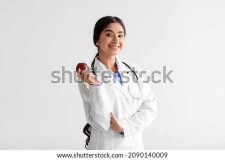 Happy pretty young indian woman doctor dentist in white coat with stethoscope hold fresh red apple on light background. Proper nutrition, vitamins, healthy diet, recommendation for perfect teeth Royalty-Free Stock Photo #2090140009