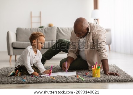 African American Little Boy And His Grandfather Sketching Together Drawing A Picture Sitting On Floor In Living Room At Home. Happy Grandpa Spending Time With His Grandchild On Weekend