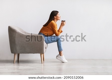 Side view of young Asian woman sitting in armchair, drinking hot aromatic coffee, relaxing against white studio wall, free space. Lovely millennial lady enjoying warm beverage on lazy day Royalty-Free Stock Photo #2090152006