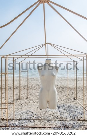 The white headless body of a mannequin hangs in a metal gazebo on the seashore in sunny weather