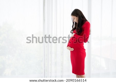 Pictures of new mothers, Europeans, Russians, pregnant Standing by the window, holding his tummy, smiling happily in the bedroom. wearing red maternity clothes