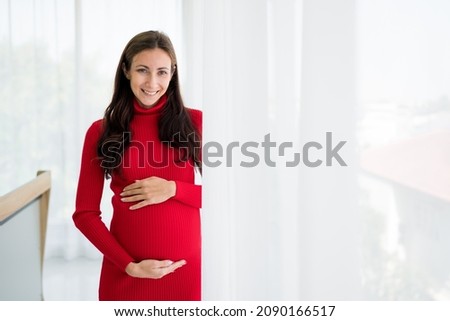 Pictures of new mothers, Europeans, Russians, pregnant Standing by the window, holding his tummy, smiling happily in the bedroom. wearing red maternity clothes