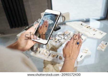 Hand of young woman trying on ring with blue gemstone and taking photograph over display with jewelry Royalty-Free Stock Photo #2090172430