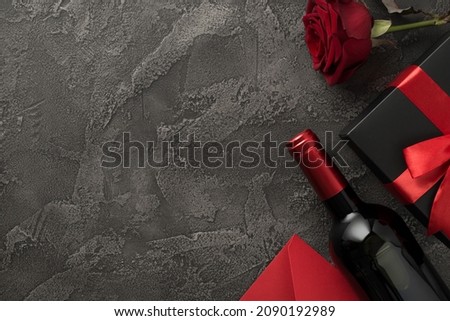 Top view photo of valentine's day decorations wine bottle black giftbox with red bow red envelope and red rose on isolated textured dark grey concrete background with blank space