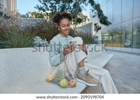 Happy relaxed curly haired woman rests after making shopping carries net bag with fruit uses smartphone dressed in fashionable clothes poses in urban setting glad to receive message from friend.