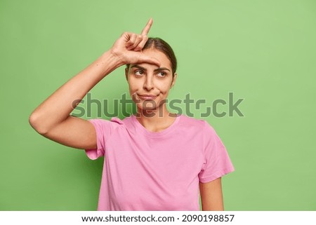 Beautiful brunette young woman does loser gesture mocks and insults someone has bad relationship with someone dressed in casual outfit isolated over vivid green background. You are outsider. Royalty-Free Stock Photo #2090198857