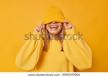 Happy young joyful woman hides eyes with hat smiles broadly shows white teeth foolishes around dressed in casual loose jumper isolated over yellow background. People and sincere emotions concept Royalty-Free Stock Photo #2090199001