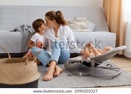 Full length portrait of happy female with dark hair and ponytail, wearing casual clothing sitting on floor with her kids, kissing her charming daughter, taking care of infant baby in rocking chair.