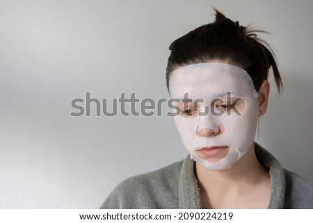 Morning beauty routine of a multicultural brunette woman with a fabric moisturizing mask with pulled hair and a gray robe on a light gray background. Concept: personal care. Take care. 