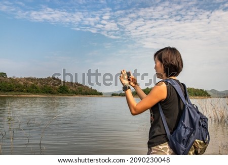 A side view of a young asia woman using her smartphone up and pointing it to take pictures view of lake shore with mountains range in background. Technology and holiday travel, No focus, specifically.