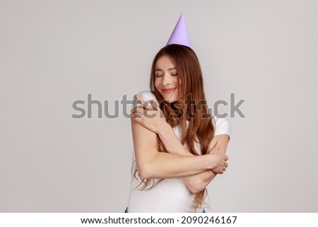 Attractive dark haired woman in party cone on head, supporting, congratulating and embracing herself on birthday, wearing white T-shirt. Indoor studio shot isolated on gray background.