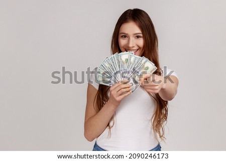 Portrait of young woman pointing to camera and holding dollar banknotes, encouraging to win lottery, earn profit. wearing white T-shirt. Indoor studio shot isolated on gray background.
