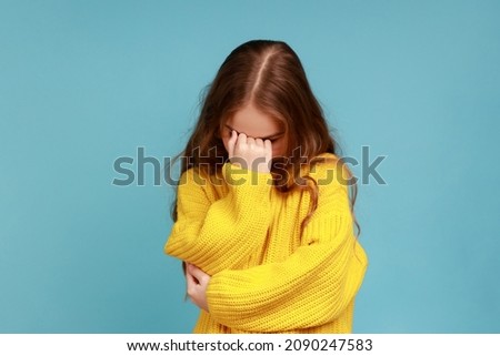 Portrait of little girl hiding face down and crying, upset about loss, feeling sorrow and regret, wearing yellow casual style sweater. Indoor studio shot isolated on blue background. Royalty-Free Stock Photo #2090247583
