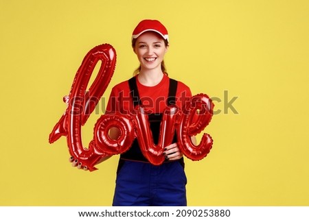 Portrait of happy smiling worker woman standing holding world love from foil balloon, looking at camera, wearing overalls and red cap. Indoor studio shot isolated on yellow background.