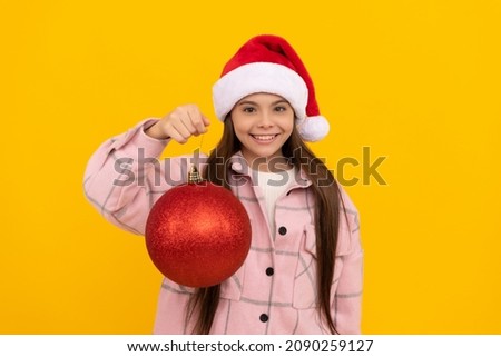 happy teen girl in santa claus hat hold christmas decorative ball on yellow background, new year