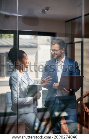Happy businessman using laptop while communicating with African American female colleague at corporate office. The view is through the glass. Royalty-Free Stock Photo #2090274040