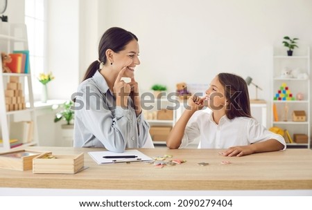 Female specialist working on kid's speech impediments. Smiling child together with professional language therapist sitting at desk in modern office, playing fun games, developing correct pronunciation Royalty-Free Stock Photo #2090279404