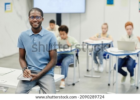 Portrait of young African-American teacher sitting on desk and smiling at camera with children in background, copy space