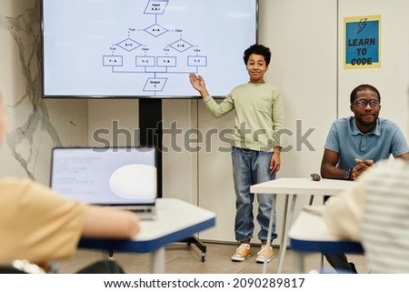 Full length portrait of smiling teenage boy giving presentation in coding class for children, copy space