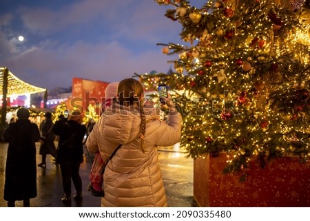 mother with little baby in arms takes photo of big shining christmas tree on night christmas market