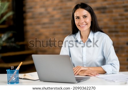 Portrait of a confident successful young adult caucasian brunette business woman, ceo, broker, manager sitting in office at table, using laptop, in formal stylish clothes, looking at camera, smiling Royalty-Free Stock Photo #2090362270