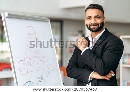 Portrait confident successful influential Indian manager or business coach in a formal suit stands at whiteboard with diagrams, in modern office, looks at camera and smiles friendly Royalty-Free Stock Photo #2090367469