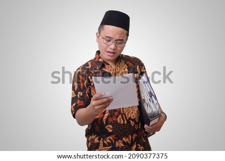 Portrait of surprised Asian man wearing batik shirt and songkok holding documents or bills with shocked facial expression. Isolated image on gray background