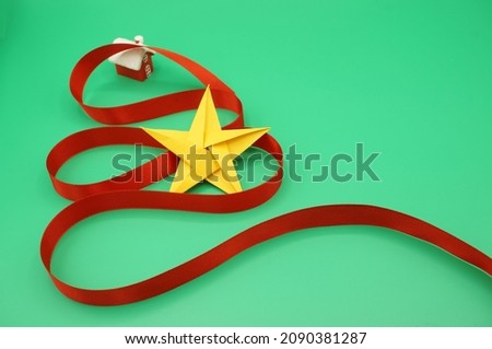 Christmas celebration theme. Green background, red ribbon around a Christmas tree in the shape of a concept, Christmas stars folded in golden paper, red hut with a snowy roof. Handmade childlike 