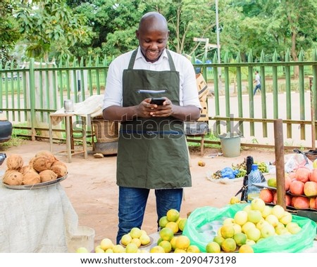 Happy African male trader or vendor holding a smart phone while standing at his fruit stall in a market place Royalty-Free Stock Photo #2090473198