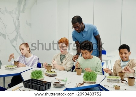 Diverse group of children planting seeds while experimenting at biology class in school with African-American teacher supervising Royalty-Free Stock Photo #2090483752