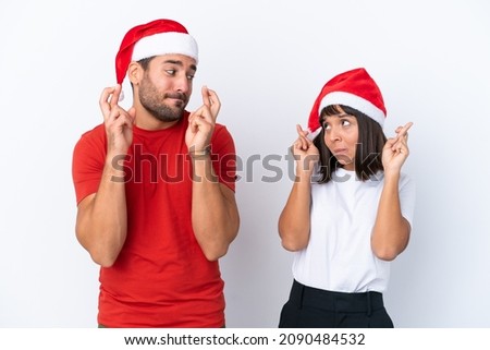 Young couple with christmas hat isolated on white background with fingers crossing and wishing the best