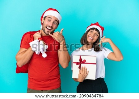 Young couple with christmas hat handing out gifts isolated on blue background making phone gesture. Call me back sign