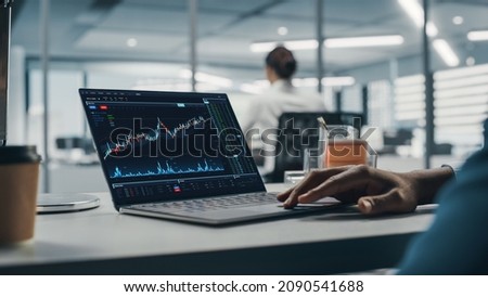 Successful Black Businessman Sitting at Desk Working on Laptop Computer in Big City Office. Hard Working Top Manager Doing Research for e-Commerce Project with Graps, Charts and Tables. Over Shoulder Royalty-Free Stock Photo #2090541688