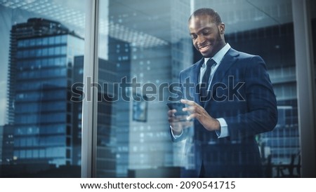 Portrait of Successful Black Businessman Wearing Suit Standing, Using Smartphone Looking out of the Window. Successful African CEO Planning e-Commerce Investment Strategy. From Outside Shot Royalty-Free Stock Photo #2090541715