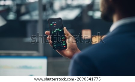 Black Businessman Holding Smartphone and Checking Stock and Cryptocurrency Market in Office. African-American Businessperson using Internet with Mobile Phone Device. Over Shoulder Shot Royalty-Free Stock Photo #2090541775