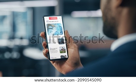 Black Businessman Using Smartphone for Checking Latest News in Office. African-American Businessperson Surfing the Internet over Mobile Phone Device. Over Shoulder Shot Royalty-Free Stock Photo #2090545090
